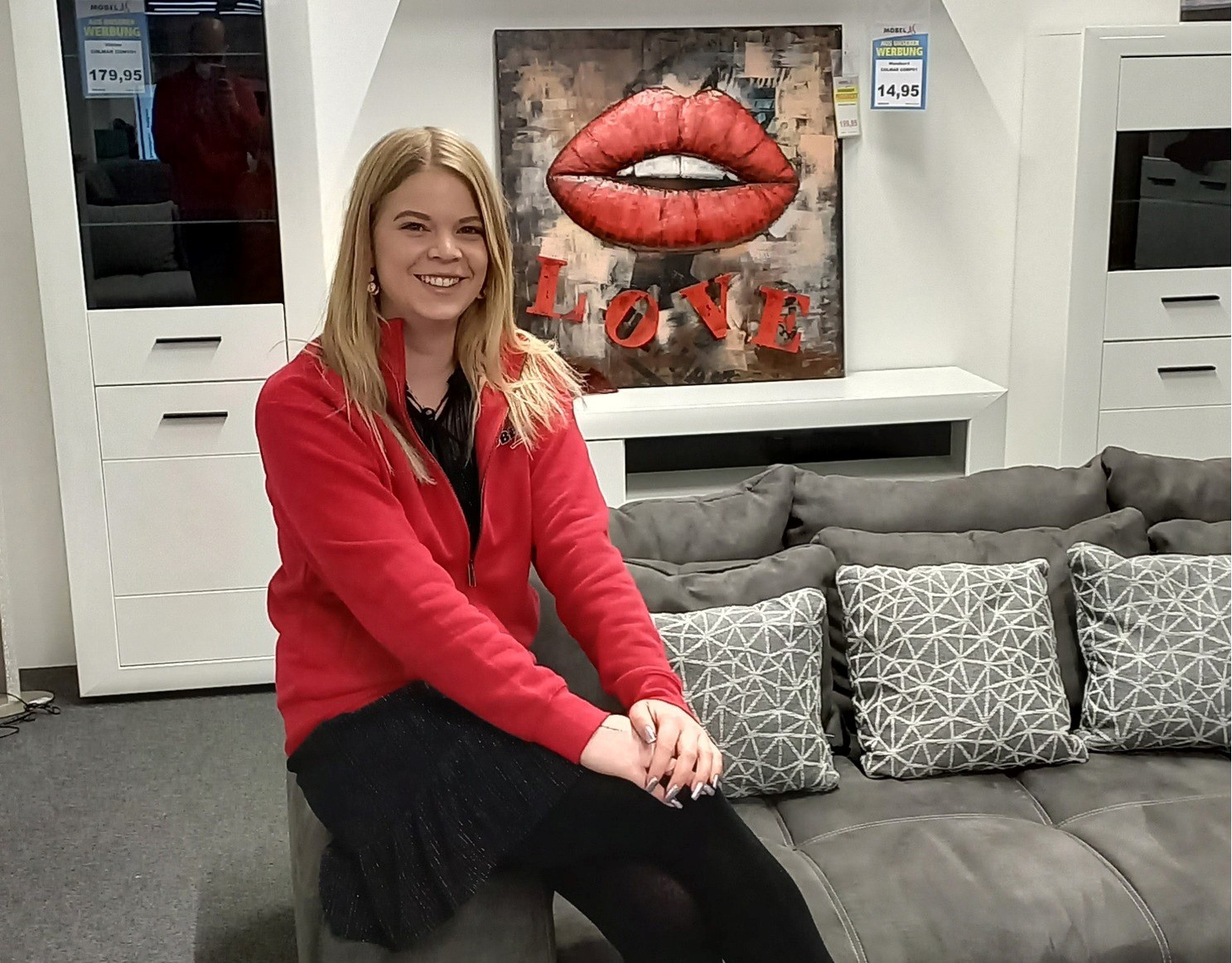 Junge Frau in roter Arbeitskleidung sitzt lächelnd auf einem grauen Sofa in einer Möbelausstellung.