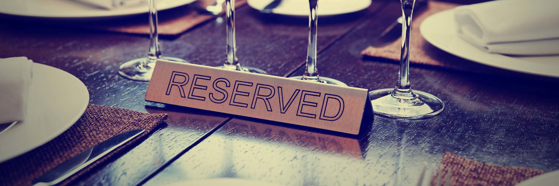 Ein elegant gedeckter Tisch in einem Restaurant ist mit weißen Tellern, Stoffservietten, Weingläsern und braunen Platzsets ausgestattet. Im Mittelpunkt steht ein Metallschild mit der Aufschrift „Reserved“.