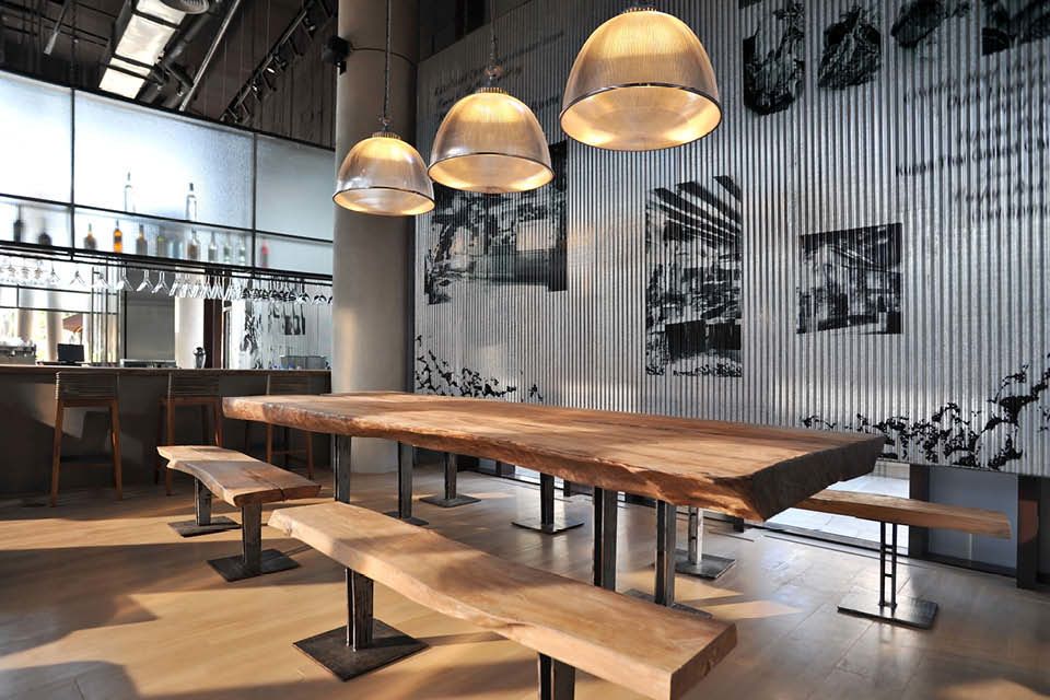 Industriell gestalteter Raum mit massiver Holztischgruppe, Pendelleuchten und Bar im Hintergrund.