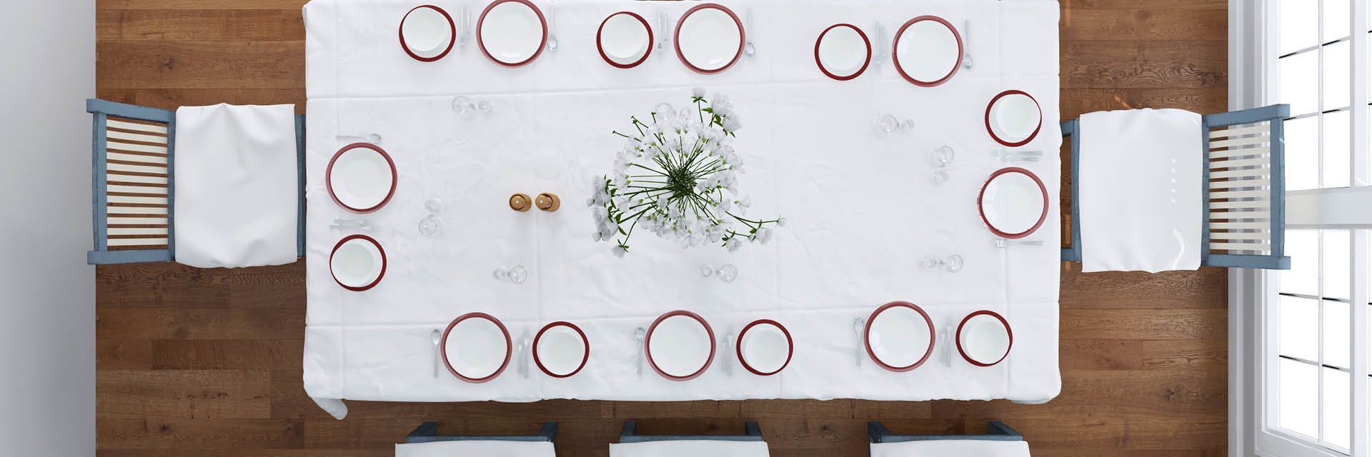 Großer, festlich gedeckter Tisch mit schlichtem weißem Tischtuch, rot umrandetem Geschirr und frischem Blumenarrangement als zentralem Blickfang.