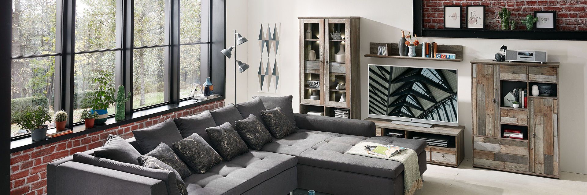 Wohnzimmer mit grauem Sofa, Pflanzen am Fenster, rustikalem TV-Möbel und Wandregalen.