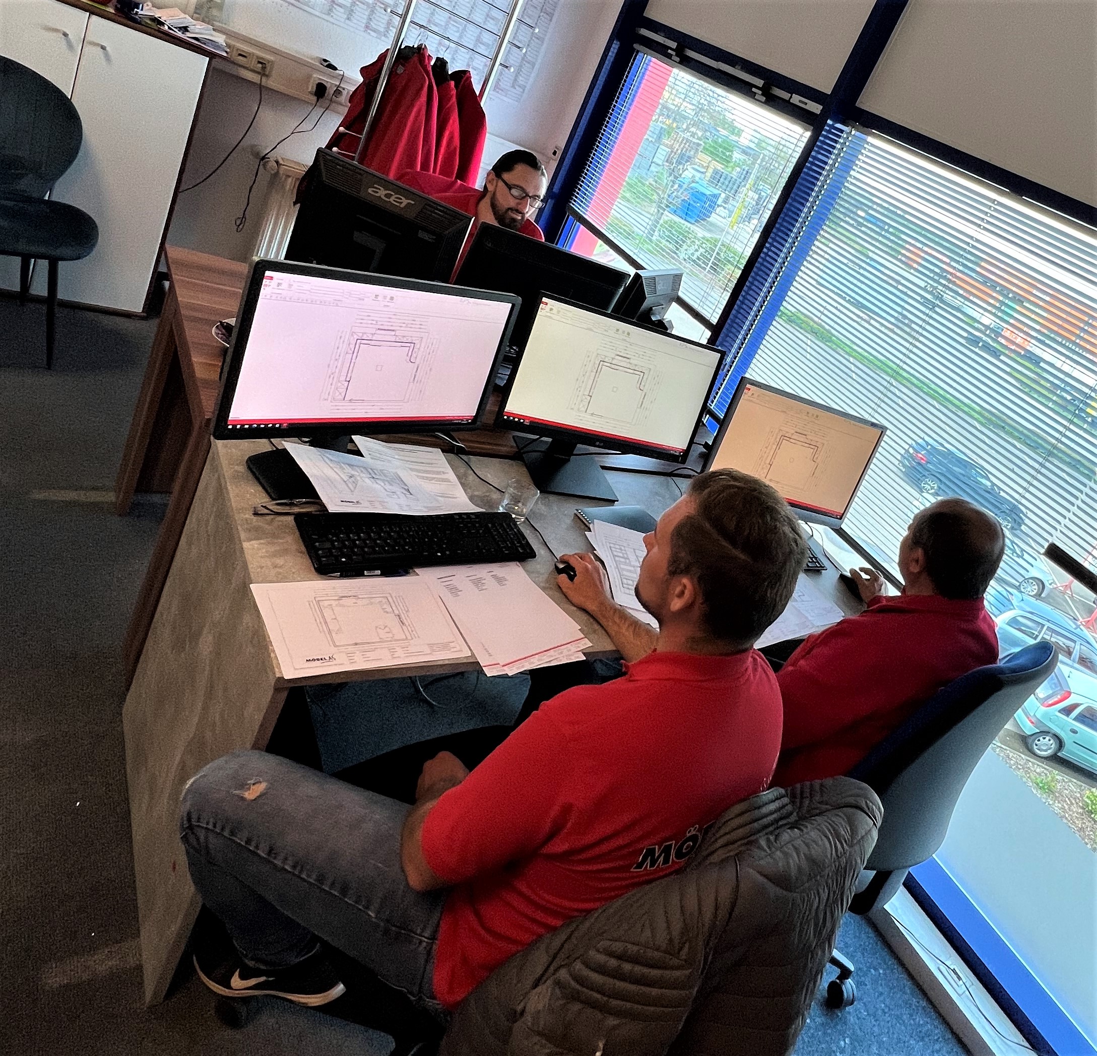 Drei Mitarbeiter in roten Möbel-AS-Shirts arbeiten an mehreren Monitoren mit Küchenplanungssoftware in einem modernen Büro mit Fensterfront.
