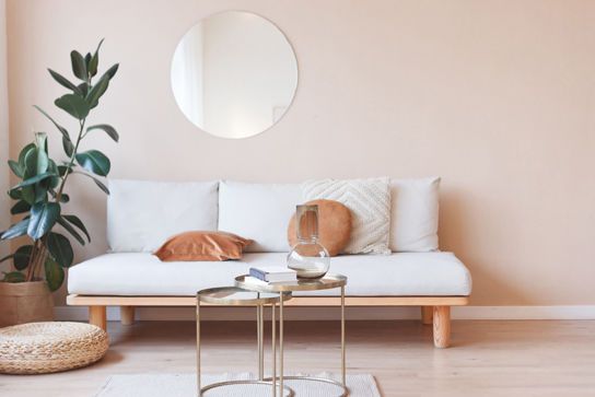 Minimalistisch eingerichtetes Wohnzimmer mit hellem Sofa, runden Couchtischen, zarten Kissen, runder Wandspiegel und großer Pflanze.