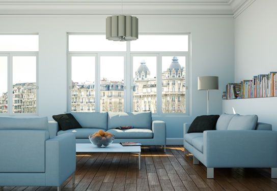 Helles Wohnzimmer mit drei hellblauen Sofas, Holzfußboden, großem Fenster mit Blick auf Altbauten und einer Bücherreihe an der Wand.