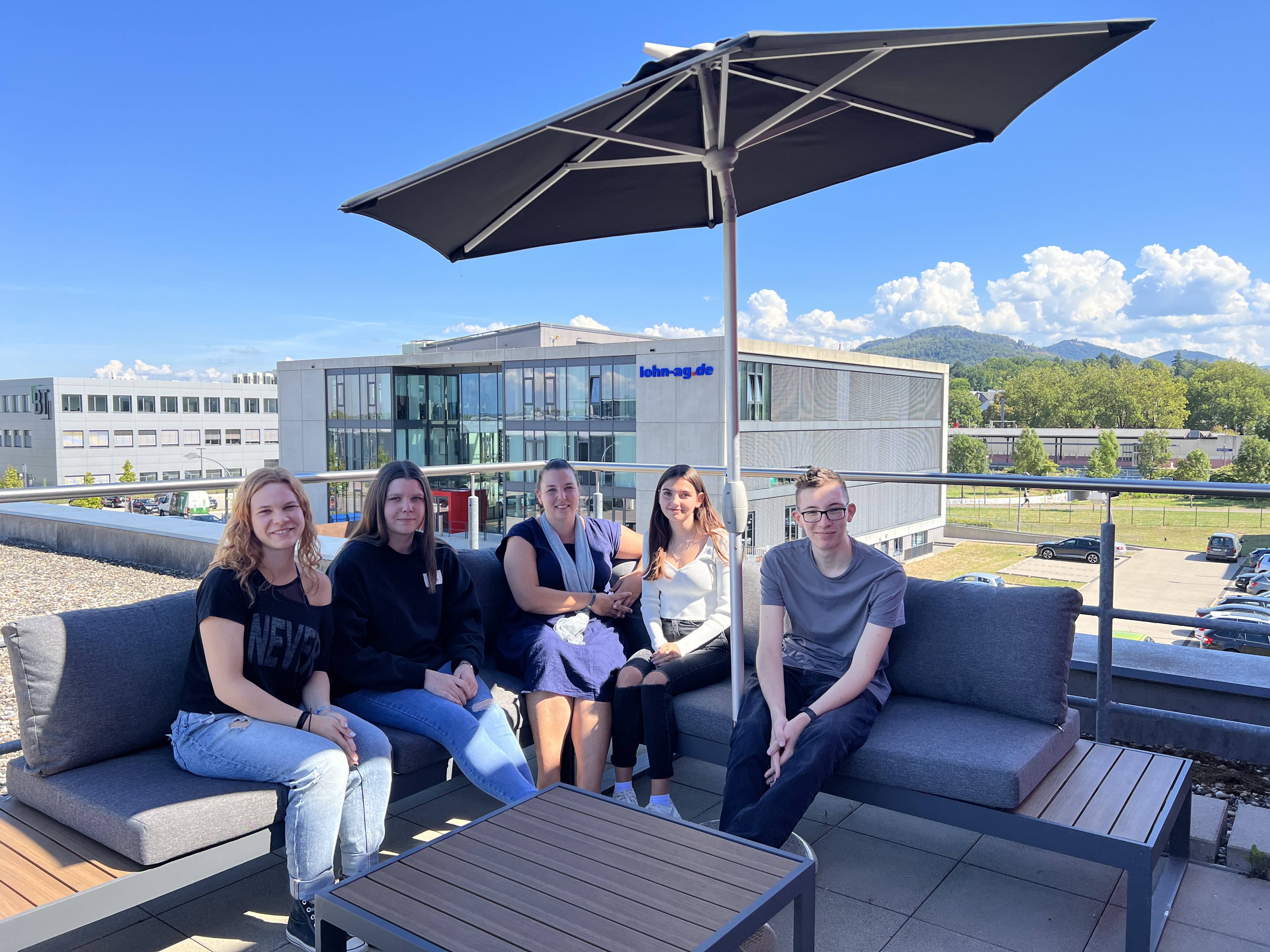 Fünf junge Erwachsene sitzen entspannt unter einem Sonnenschirm auf einer Dachterrasse mit Blick auf ein Bürogebäude und grüne Hügel im Hintergrund.