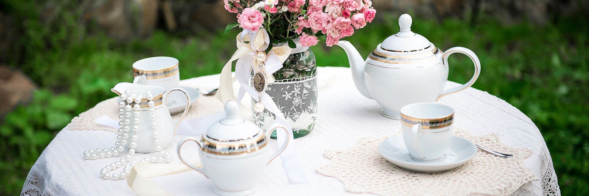 Liebevoll gedeckter Gartentisch mit edlem Porzellanservice, goldenen Verzierungen, frischen Blumen und nostalgischer Spitze – perfekt für eine stilvolle Teezeit im Grünen.