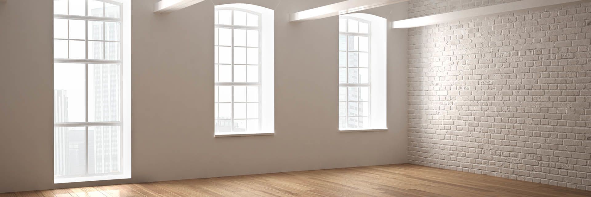 Leerer, heller Raum mit großen Sprossenfenstern, weißer Ziegelwand und Holzfußboden.