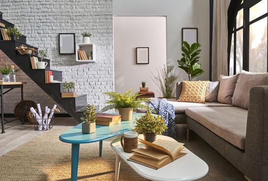 Gemütliches Wohnzimmer mit vielen Zimmerpflanzen, moderner Treppe, heller Eckcouch und bunten Tischen mit Büchern und Tasse.