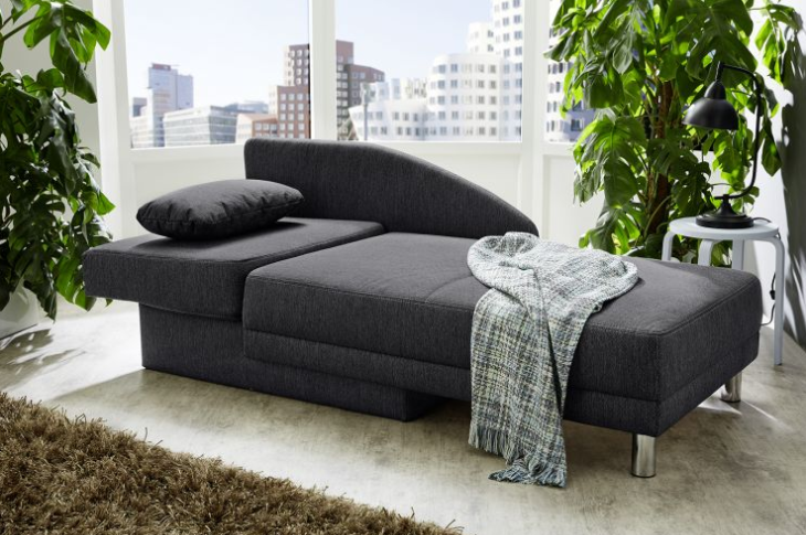 Dunkelgraues Sofa mit verstellbarer Rückenlehne, Kissen und Decke, in hellem Raum mit Pflanzen und Stadtblick.