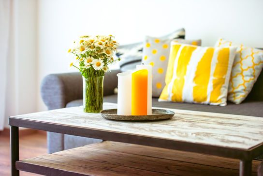 Holztisch mit einer Vase mit Blumen und einer Kerze, im Hintergrund ein graues Sofa mit gelben Kissen.